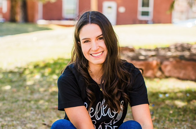 Katee Hurst smiles as she sits on the grass on NAU's campus with an apple-shaped sign that says "Miss Hurst."