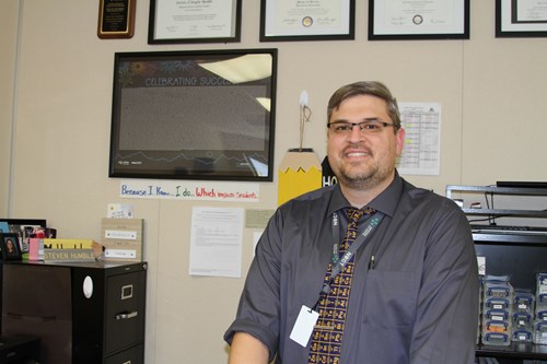 Science teacher Steven Humble, NBCT stands by his desk in his classroom, smiling and looking at the classroom.