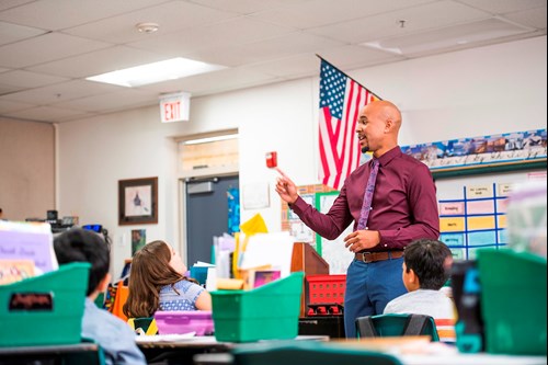 A new teacher stands in front of a class of elementary students, smiling as he speaks to them.