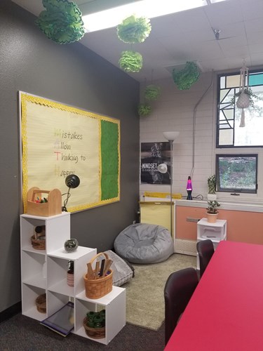 An image of a corner of Winiesdorffer's classroom that includes a beanbag, a couple floor pillows on top of a rug, a few lamps, several baskets holding school supplies, a plant, fluffy-looking green paper decorations hanging from the ceiling, and a poster that reads "Mistakes Allow Thing to Happen," the first letters highlighted in different colors to highlight that they spell "MATH."