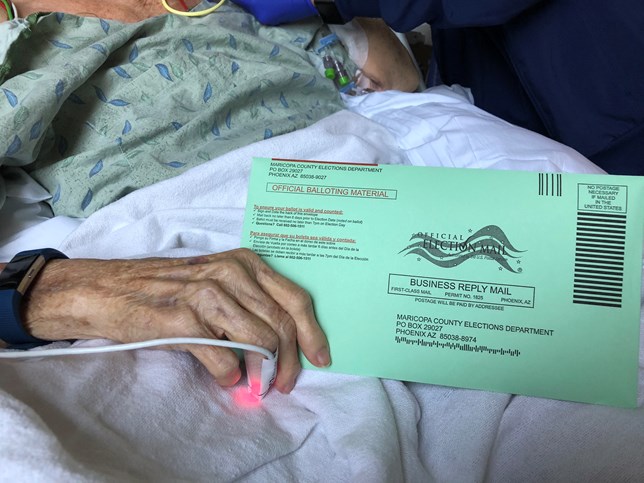 The hand of a woman in the ICU holds a green Maricopa County ballot envelope.