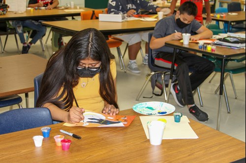 A fifth-grade girl adds white to a Halloween-themed painting of a moon, a yellow and orange sky, and a black castle, on her desk.
