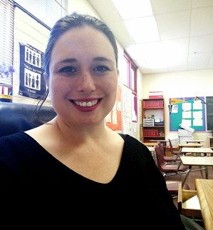 Teacher Sarah Garcia smiles at the camera while seated at her desk. We see a few students' desks, bookshelves, and bulletin boards over her shoulder.