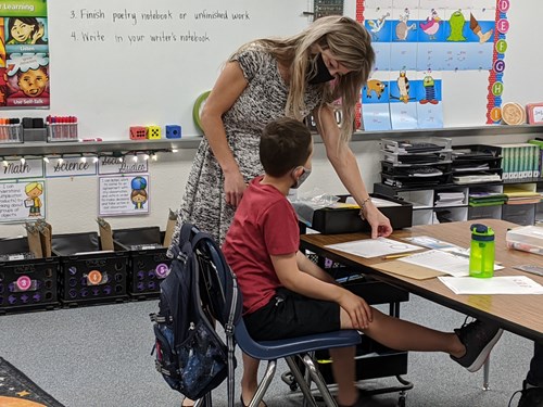 Third-grade teacher Sheri Ramirez, in a mask, speaks to a student, also in a mask, seated as his desk.