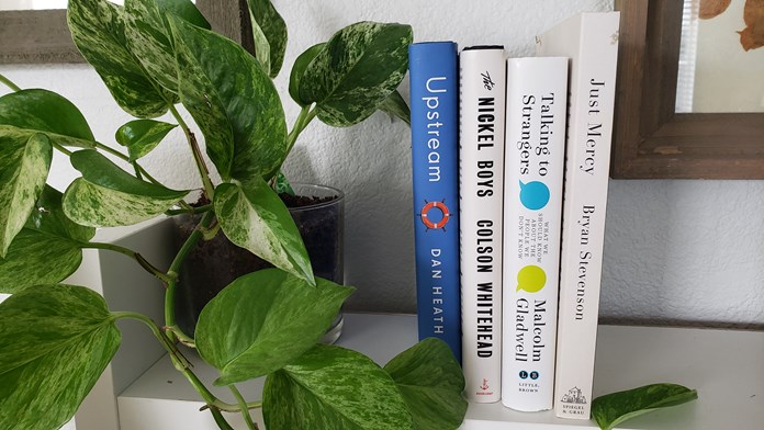 Next to a plant on a book shelf sit four books: Dan Heath's Upstream, Colson Whitehead's The Nickel Boys, Malcolm Gladwell's Talking to Strangers, and Bryan Stevenson's Just Mercy.